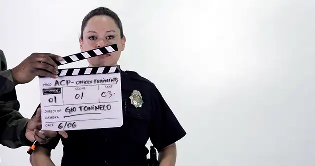 A female police officer stands behind a movie slate on a white studio.
